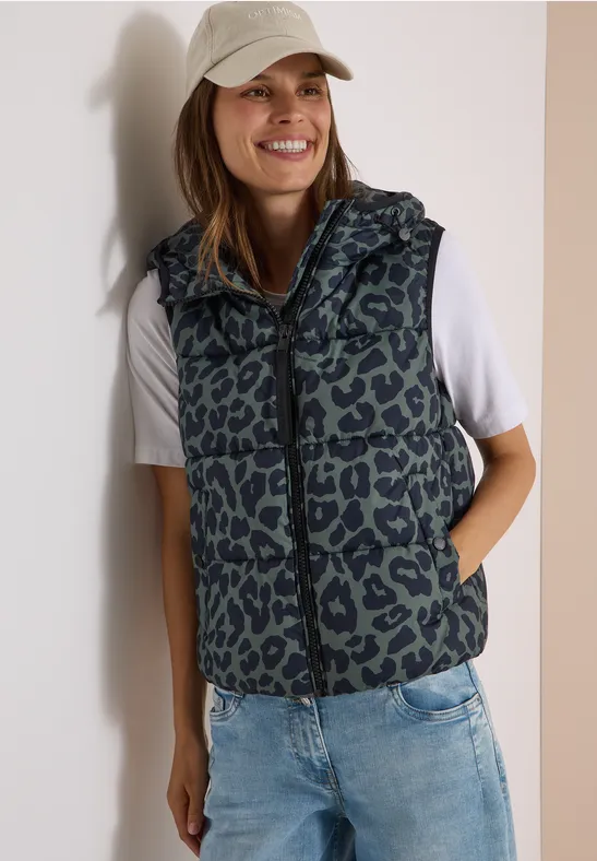 Junge Frau trägt Jeans, weißes T-Shirt, Leopard-Weste und Baseballcap, lächelt.