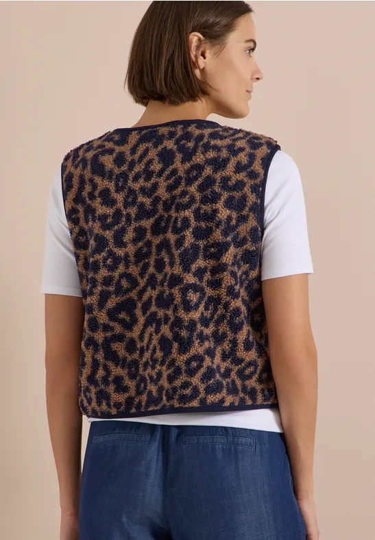 Frau seitlich in weißem T-Shirt, braun-gemusterter Leoparden-Weste und Jeanshose vor beigem Hintergrund.