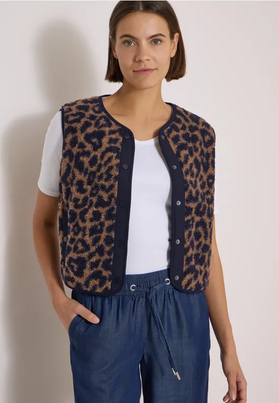 Frau trägt weißes Shirt, leo-Strickweste ohne Ärmel und blaue Hose vor hellgrauem Hintergrund.
