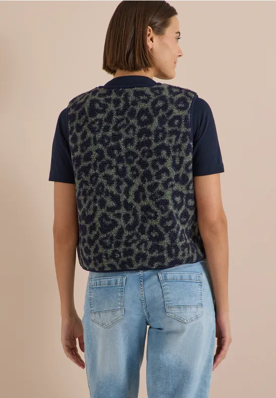 Rückenansicht einer Frau in dunkelgrün-schwarzer Leo-Weste über schwarzem Shirt und hellblauen Jeans.