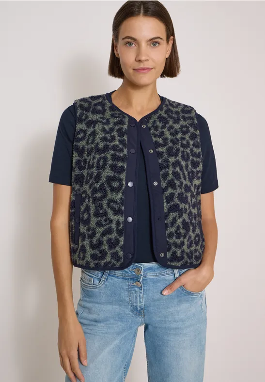 Frau in blauem T-Shirt mit grüner, leo-gemusterter Weste und hellen Jeans vor weißem Hintergrund.