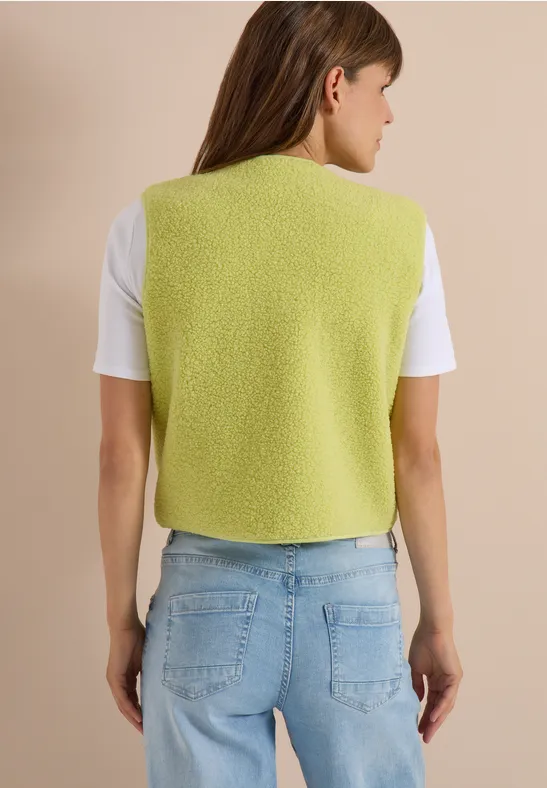 Frau von hinten in hellgrüner Strickweste, weißem T-Shirt und blauen Jeans vor neutralem Hintergrund