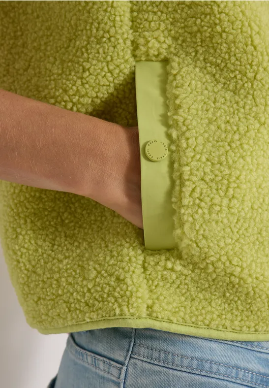 Hand steckt in Tasche einer hellgrünen Teddy-Fleecejacke, darunter hellblaue Jeans.
