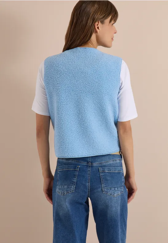 Frau von hinten in hellblauer Teddy-Weste, weißem T-Shirt und blauen Jeans vor beigem Hintergrund.