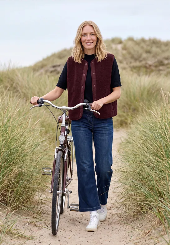 Frau schiebt Fahrrad auf Dünenpfad zwischen hohem Gras am Strand.