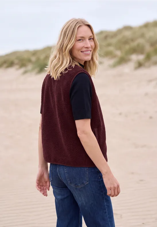Eine lächelnde Frau in einer bordeauxfarbenen Strickweste und Jeans steht am Strand und blickt über die Schulter.