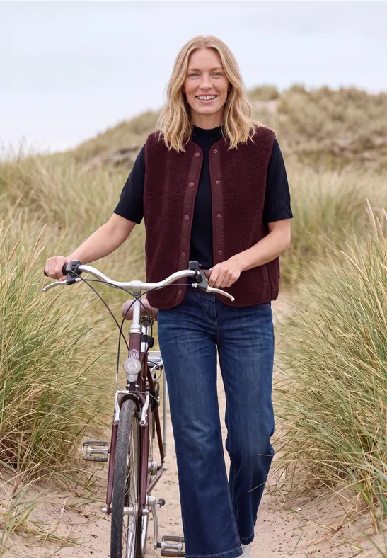 Eine lächelnde Frau geht mit einem Fahrrad durch grasbewachsene Dünen am Strand.
