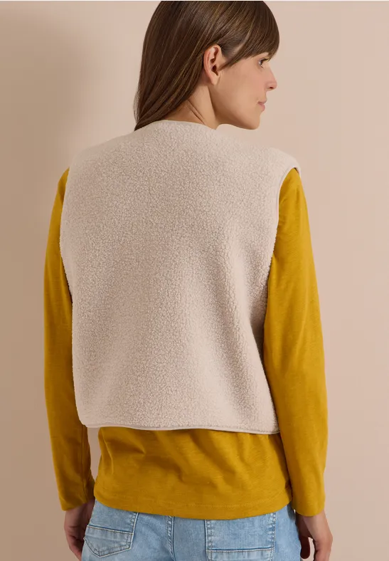 Frau von hinten mit cremefarbener Teddyweste über senfgelbem Langarmshirt und Jeans.