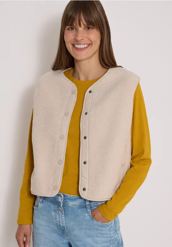Lächelnde Frau trägt gelbes Shirt und beige Weste, lässig in Jeans, Hände in Tasche.