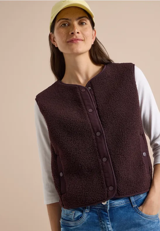 Frau mit dunkelbrauner Fleeceweste, weißem Langarmshirt, Jeans und beiger Mütze.