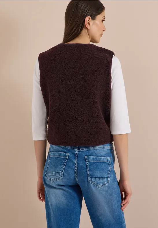 Rückansicht einer Frau in weißem Shirt, lila Bouclé-Weste und blauen Jeans vor beigem Hintergrund.