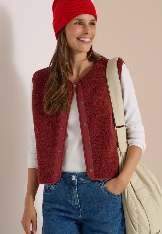Eine junge Frau trägt rote Weste und Mütze, ein weißes Shirt, Jeans und eine Cremetasche, sie lächelt.