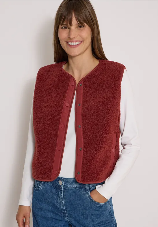 Frau mit roten Strickweste, weißem Langarmshirt und Jeans, Hände in den Taschen, lächelnd vor hellem Hintergrund.