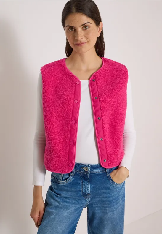 Eine Frau trägt eine pinke Weste über einem weißen Langarmshirt und blauen Jeans.