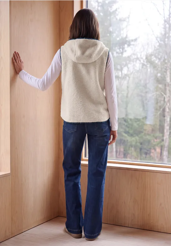 Person in Fleece-Weste und Jeans steht in einem Holzraum vor grossem Fenster und blickt in den Wald.