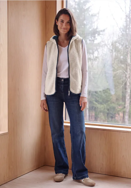 Eine Frau steht lässig in heller Fleecejacke und Jeans vor einem großen Fenster mit Blick auf den Wald.