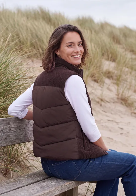 Glimlachende vrouw in bruine bodywarmer zit op een bankje in de duinen