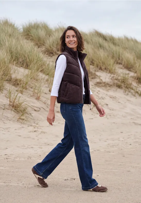 Vrouw in casual outfit wandelt over het zandstrand met duinen en helmgras op de achtergrond.