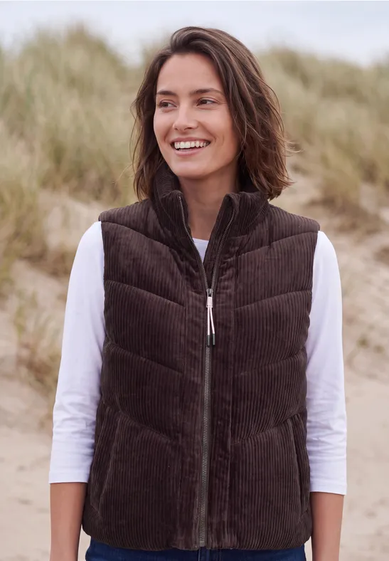 Een vrouw glimlacht bij zandduinen aan zee, ze draagt een bruine corduroy bodywarmer en een wit shirt met lange mouwen.