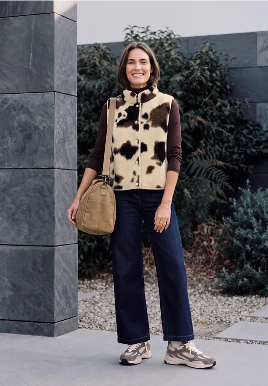 Frau steht vor modernem Gebäude mit Kuhflecken-Weste, braunem Pullover, blauen Hosen, Sneakers und Tasche.