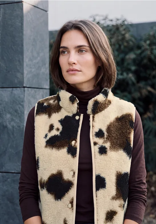 Frau mit kurzem Bob trägt beige-braune Kuhfell-Weste über dunklem Rollkragenpullover vor moderner Fassade.