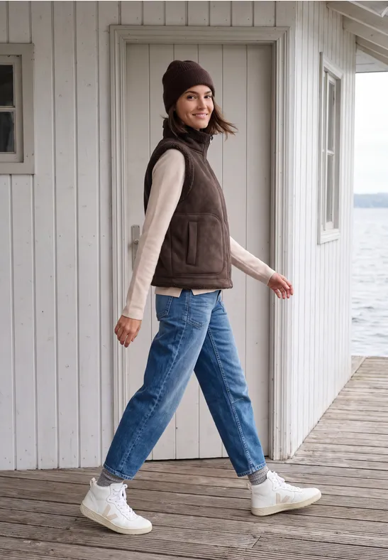 Eine junge Frau spaziert auf einem Holzsteg am See, trägt braune Weste, Beanie, Jeans und weiße Sneakers.