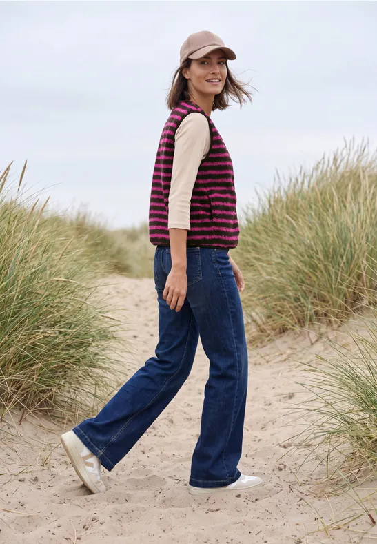 Frau in Jeans, gestreiftem Pullunder und Mütze geht am Strand durch Dünengras.