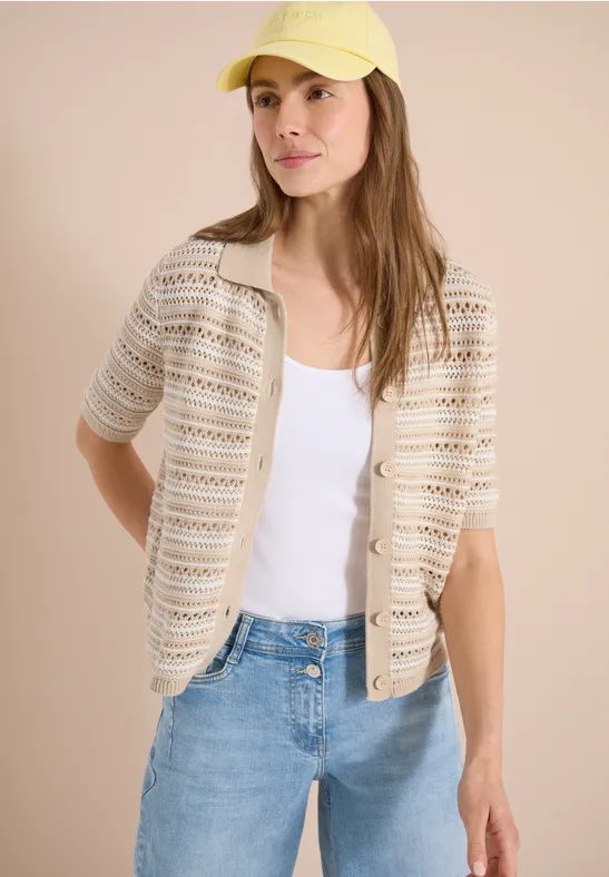 Frau trägt gehäkelte beige Strickjacke über weißem Top, Jeans und gelber Kappe.