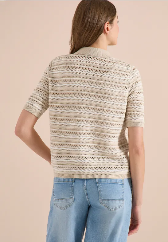 Rückansicht einer Frau in beige gemustertem Strick-T-Shirt mit kurzen Ärmeln und blauen Jeans vor hellem Hintergrund.