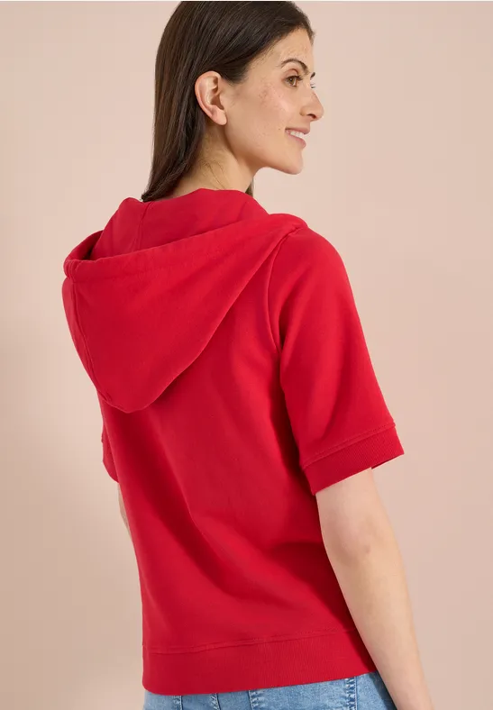 Achteraanzicht van een vrouw in een rood hoodietje met korte mouwen voor een beige achtergrond.