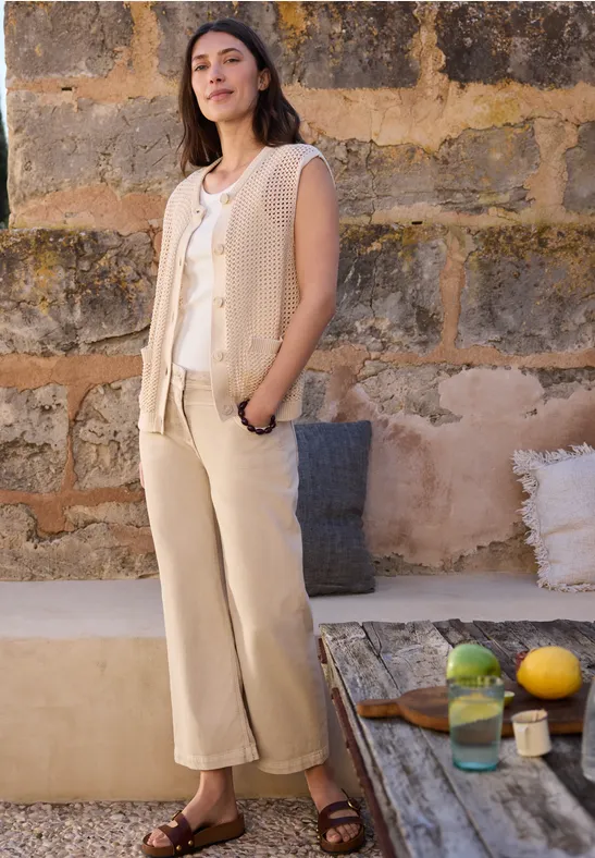 Frau in beiger Strickweste, weißem Top und beigen Hosen steht vor einer Steinmauer neben Tisch mit Obst und einem Glas.