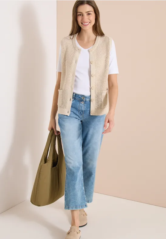 Frau trägt weißes T-Shirt, beige Weste, blaue Jeans, olivgrüne Tasche und beige Schuhe vor neutralem Hintergrund.