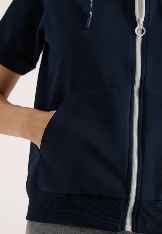 Eine Person trägt einen dunkelblauen Kapuzenpullover mit Reißverschluss und steckt die Hand in die Fronttasche.