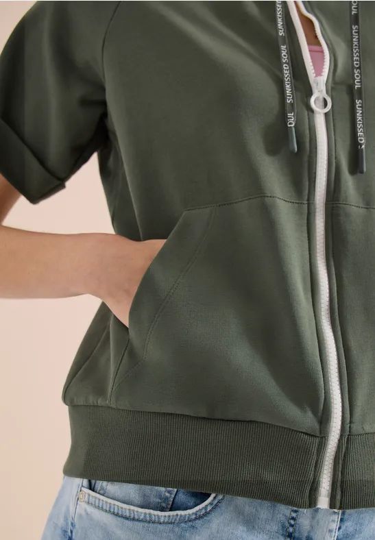 Olivegrüne, kurzärmlige Kapuzenjacke mit Reißverschluss und Taschen, kombiniert mit Jeans.