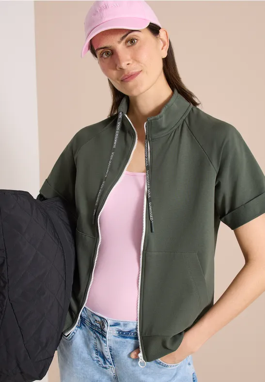 Frau in grüner Kurzarmjacke, rosafarbenem Top und Jeans, hält eine schwarze gesteppte Jacke.