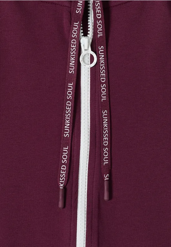 Nahaufnahme eines bordeauxfarbenen Hoodies mit weißem Reißverschluss und Kordeln mit Aufdruck SUNKISSED SOUL
