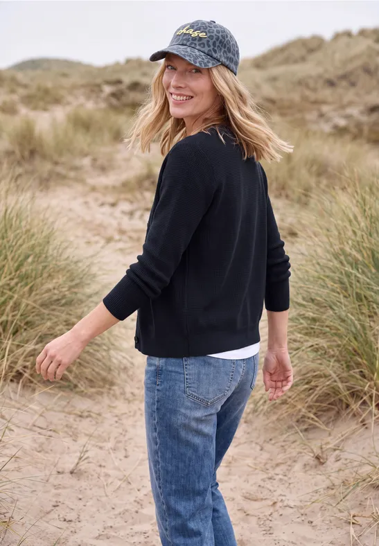 Eine lächelnde Frau in Jeans und Pullover spaziert am Dünenstrand, schaut über die Schulter in die Kamera.