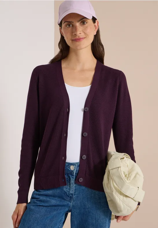 Frau in lila Strickjacke, weißem Top und Jeans hält eine helle, gesteppte Tasche.