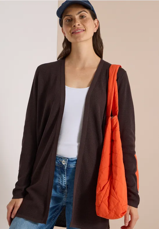 Frau in Jeans, weißem Top, dunkler Strickjacke, blauer Mütze und orangefarbener Tasche