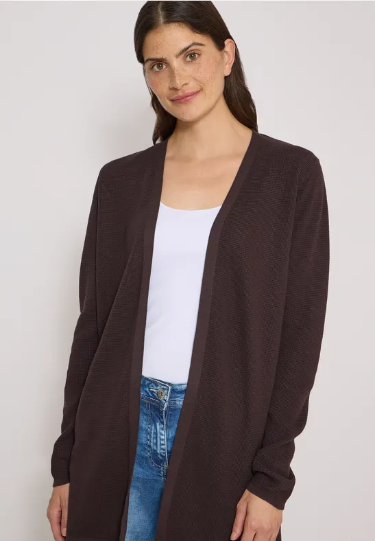 Schlanke Frau mit brauner Strickjacke, weißem Shirt und Jeans vor hellem Hintergrund.