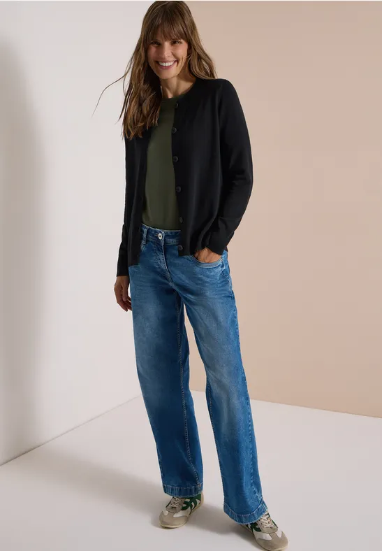 Eine Frau steht in Jeans, grünem Shirt und schwarzer Strickjacke, lächelnd.