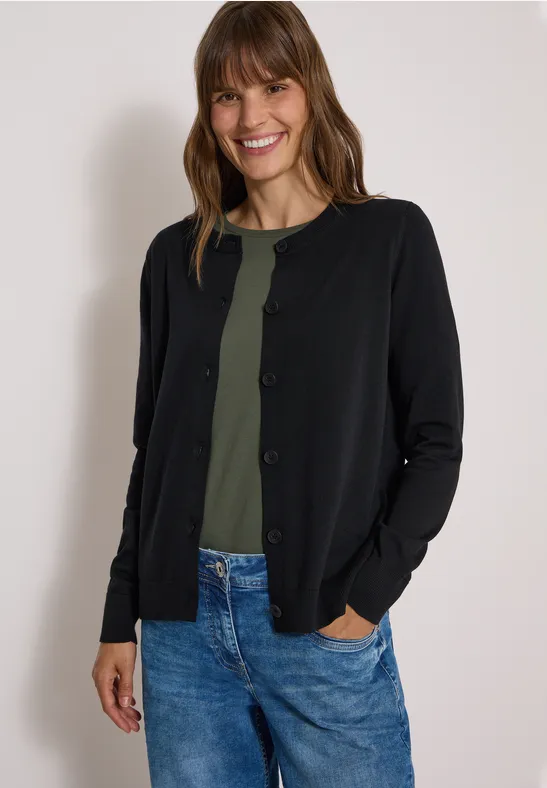 Eine lächelnde Frau in schwarzer Strickjacke, grünem Shirt und Jeans, Hände in den Taschen.