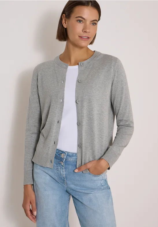 Eine Frau mit kurzem braunen Haar trägt ein graues Knopf-Cardigan über weißem Oberteil und blauen Jeans.