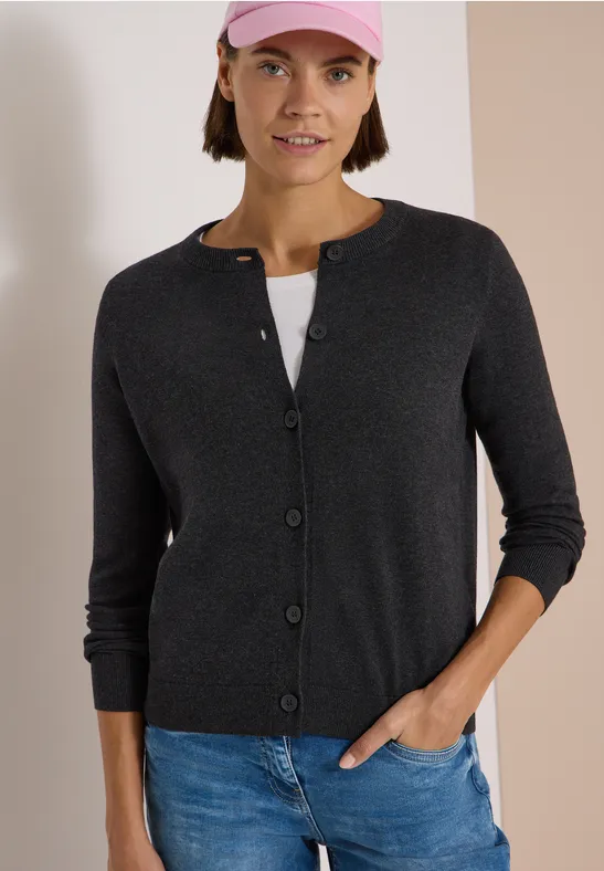 Frau in grauer Strickjacke, Jeans und rosa Cap vor schlichtem zweifarbigem Hintergrund.