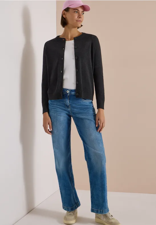 Eine Frau in schwarzer Strickjacke, weißem Top, blauen Jeans und rosa Basecap steht an einer Ecke.