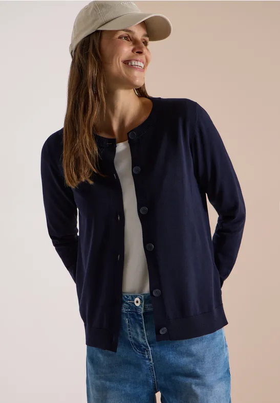Frau in Jeans, weißem Top, dunkelblauer Strickjacke und Cap lächelt vor hellem Hintergrund.