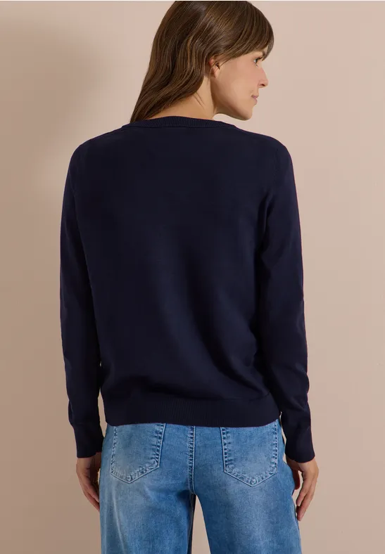 Rückansicht einer Frau mit braunen Haaren in dunkelblauem Pullover und hellblauen Jeans vor beigem Hintergrund.