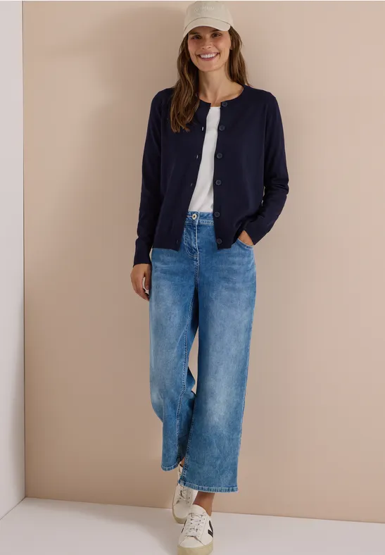 Frau in weitem Jeans, weißem Shirt, dunkler Strickjacke, Sneakern und Cap vor beigem Hintergrund.