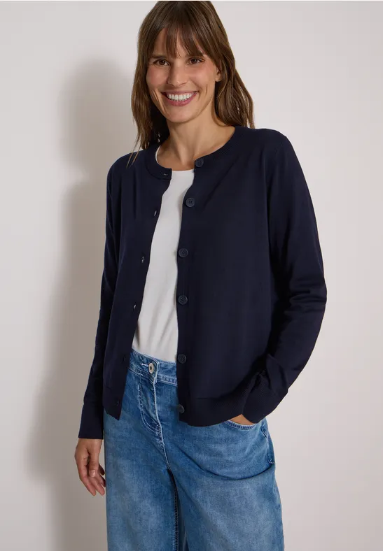 Eine lächelnde Frau trägt eine dunkelblaue Strickjacke, weißes Shirt und Jeans.