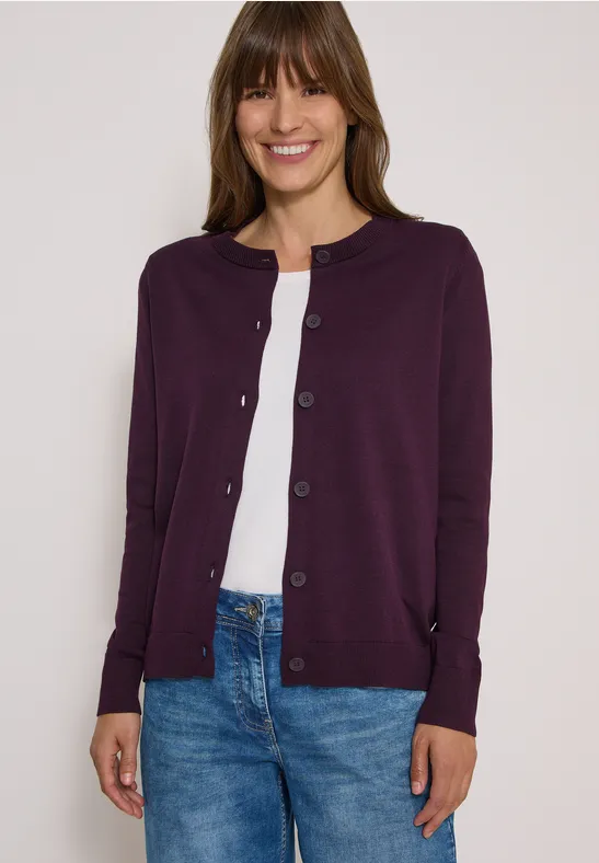Frau in lila Strickjacke, weißem Shirt und blauen Jeans lächelt.